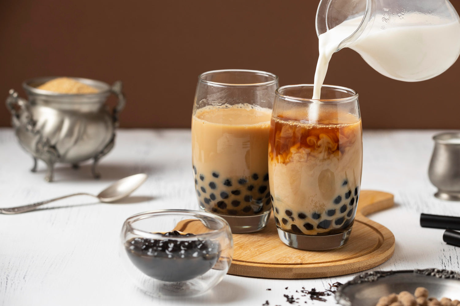 Two glasses of bubble tea with milk being poured into one, on a table with a brown background.